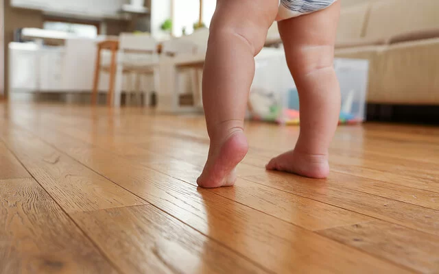 Nahaufnahme der nackten Beine eines gewindelten Kleinkindes, das auf einem Holzfußboden die ersten Gehversuche macht.