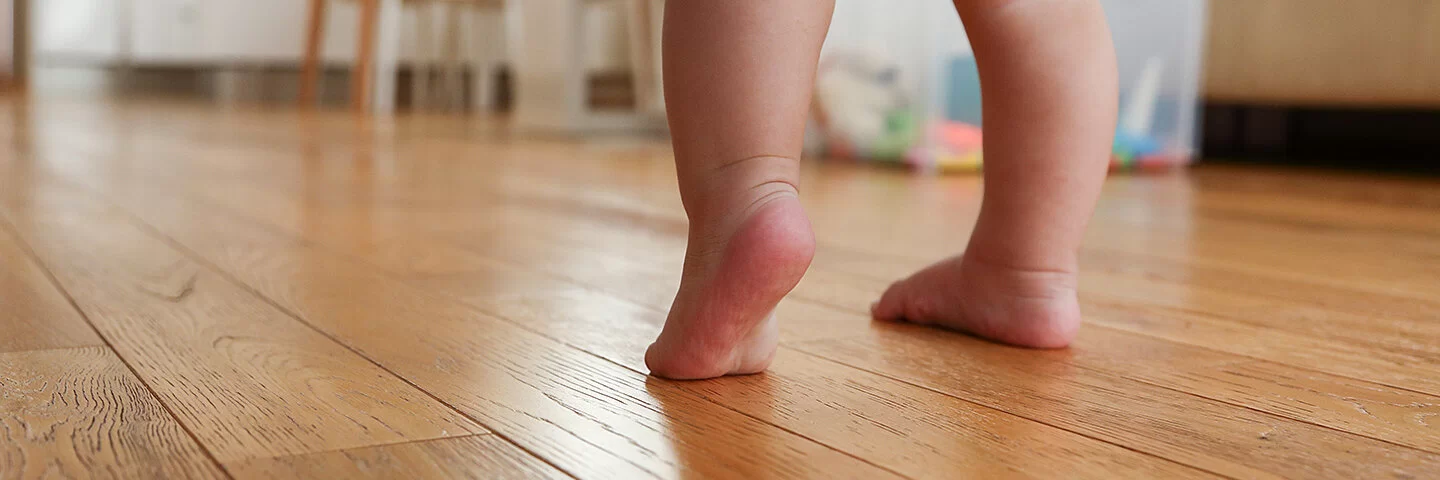 Nahaufnahme der nackten Beine eines gewindelten Kleinkindes, das auf einem Holzfußboden die ersten Gehversuche macht.