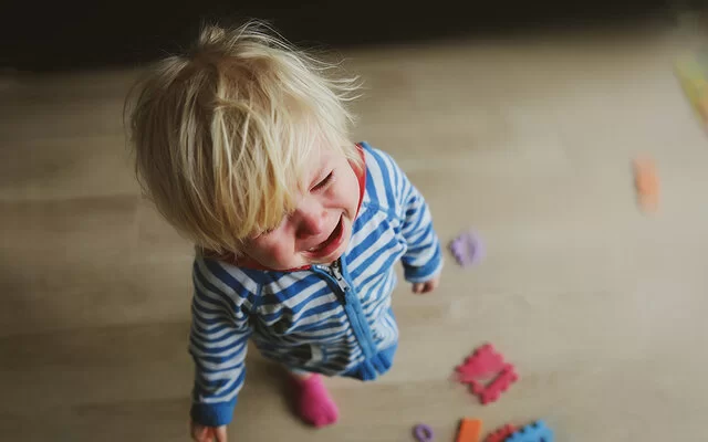 Nahaufnahme von oben: Ein weinendes blondes Kleinkind steht in einem gestreiften Strampler auf einem Holzfußboden. Zu seinen Füßen liegt verstreutes Spielzeug.