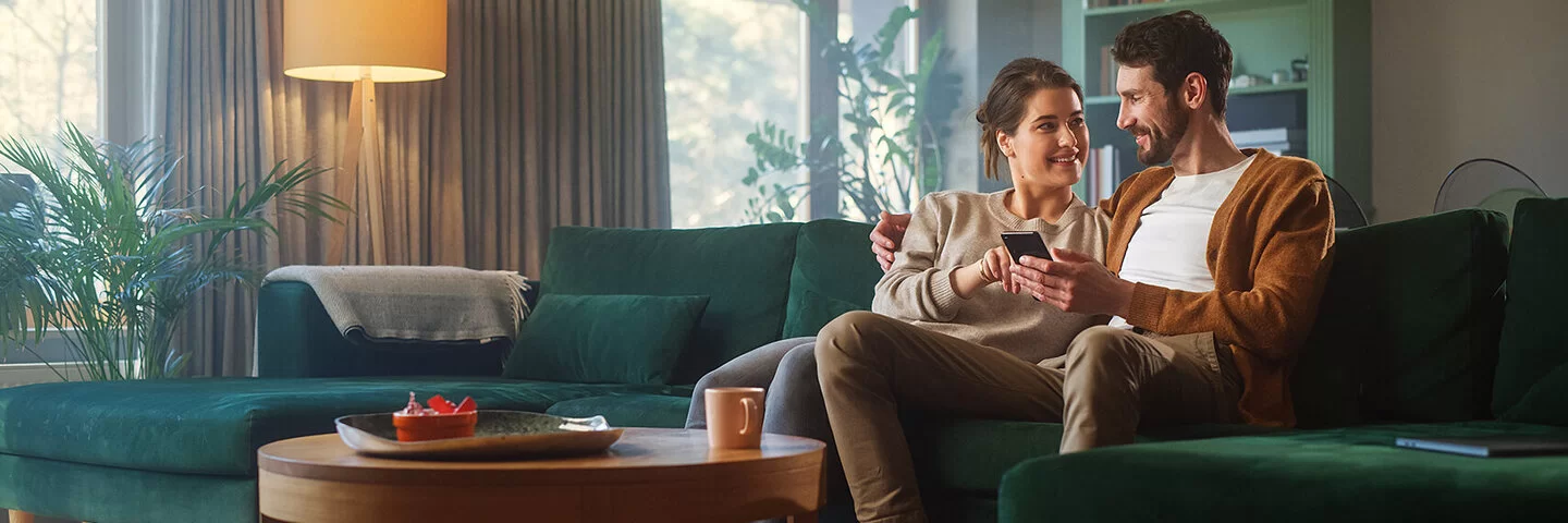 Ein Pärchen in den Dreißigern sitzen nah beieinander auf dem Sofa und lächeln sich an. Der Mann hält ein Smartphone in seiner linken Hand, die Frau zeigt mit ihrer rechten Hand auf das Display.