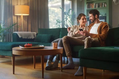 Ein Pärchen in den Dreißigern sitzen nah beieinander auf dem Sofa und lächeln sich an. Der Mann hält ein Smartphone in seiner linken Hand, die Frau zeigt mit ihrer rechten Hand auf das Display.