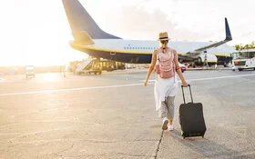 Eine junge Frau in sommerlicher Kleidung geht am Flughafen mit einem Rollkoffer und einem Rucksack auf ein Flugzeug zu.