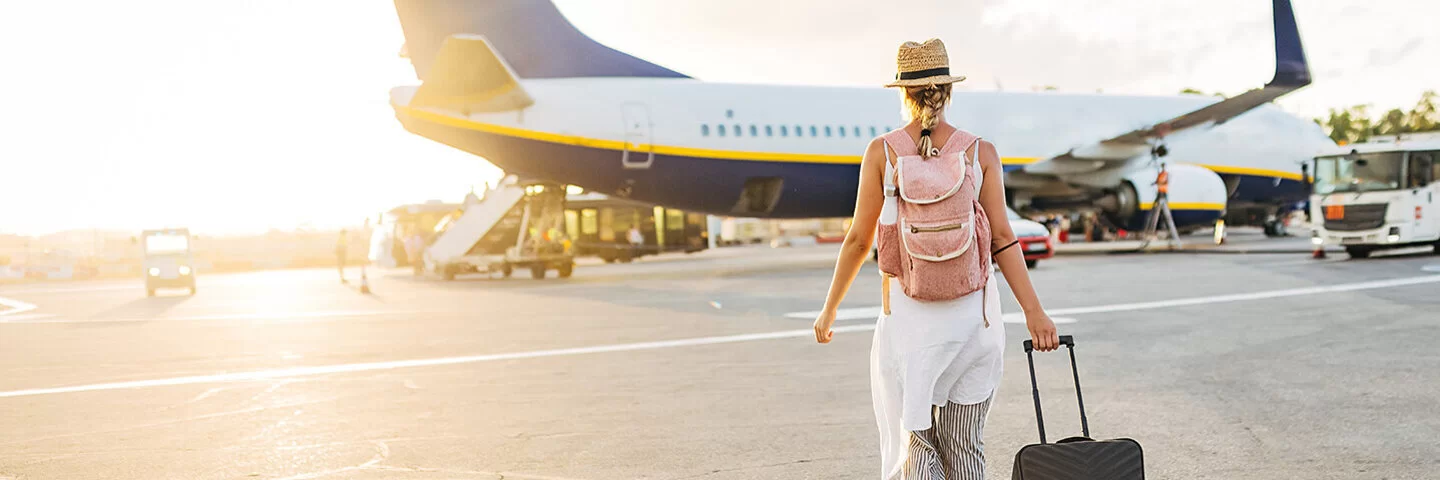 Eine junge Frau in sommerlicher Kleidung geht am Flughafen mit einem Rollkoffer und einem Rucksack auf ein Flugzeug zu.