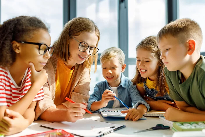 Kinder sitzen gemeinsam an einem Tisch im Klassenzimmer. Eine Lehrkraft sitz dabei und hilft bei den Hausaufgaben.