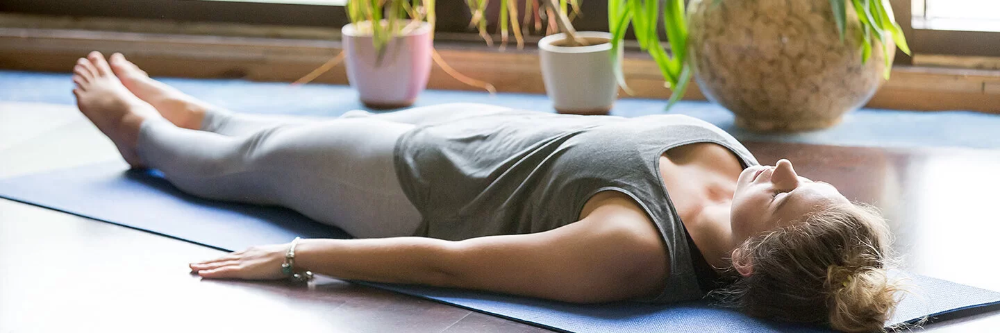 Eine Frau in Sportkleidung liegt mit dem Rücken auf einer Yogamatte und hat die Augen geschlossen. Im Hintergrund zu sehen sind Topfpflanzen und bodentiefe Fenster.