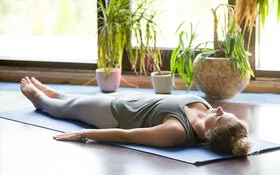 Eine Frau in Sportkleidung liegt mit dem Rücken auf einer Yogamatte und hat die Augen geschlossen. Im Hintergrund zu sehen sind Topfpflanzen und bodentiefe Fenster.