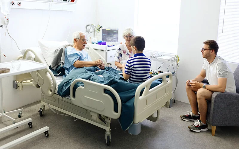 Ein älterer Patient liegt in einem Krankenzimmer und hat Besuch von einer älteren Dame, einem jungen Mann und einem Jungen, die neben dem Bett sitzen.