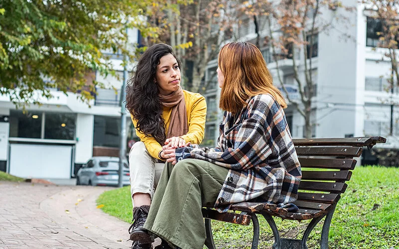 Zwei junge Frauen sitzen sich auf einer Parkbank in einer Grünanlage vor einem weißen Apartmentgebäude gegenüber. Sie scheinen in ein ernstes Gespräch vertieft. Die rechts sitzende Frau hat das Gesicht der Kamera zugewandt. Sie hat lange schwarze Haare und trägt einen gelben Pullover, beigefarbene Hosen und einen braunen dünnen Schal. Die links sitzende Frau ist der Kamera abgewandt. Sie trägt ein großes kariertes Flanellhemd als Jacke und eine weite grüne Hose.