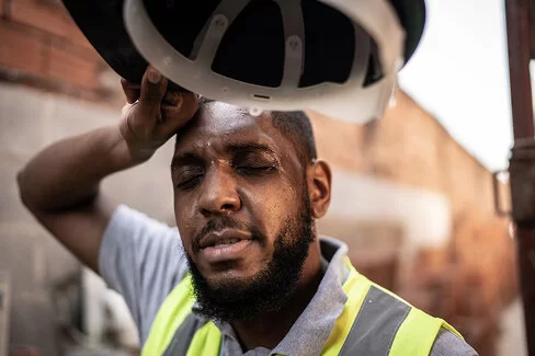 Ein Bauarbeiter mit hellgrauem Shirt und gelber Warnweste hat seinen Helm in der rechten Hand und wischt sich mit dem Handrücken über die Stirn.