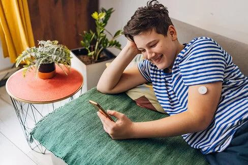 Ein Junge im Teenageralter liegt entspannt auf einem Sofa und schaut lächelnd auf sein Smartphone. Auf seinem linken Arm trägt er einen Glukosesensor.