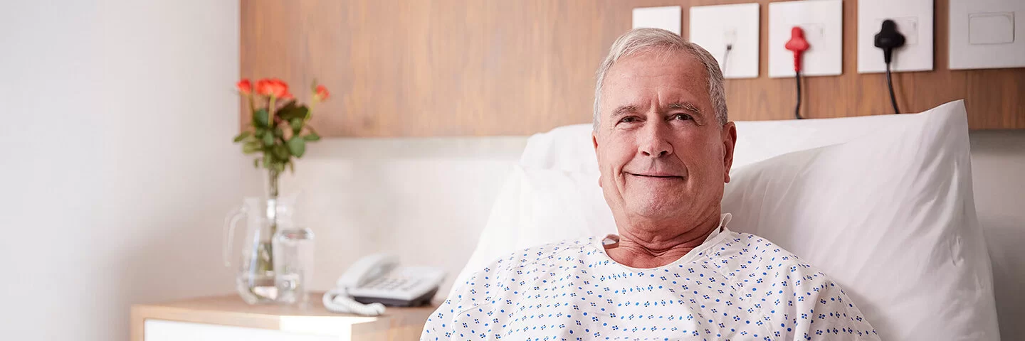 Ein älterer Mann sitzt aufrecht in einem Krankenbett, seine Hände liegen gefaltet auf seinem Bauch. Im Hintergrund sind ein Telefon und Blumen in einer Vase zu sehen.