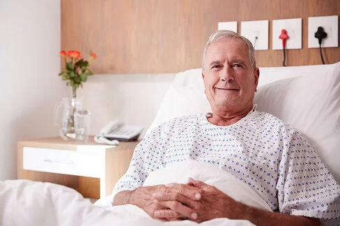 Ein älterer Mann sitzt aufrecht in einem Krankenbett, seine Hände liegen gefaltet auf seinem Bauch. Im Hintergrund sind ein Telefon und Blumen in einer Vase zu sehen.