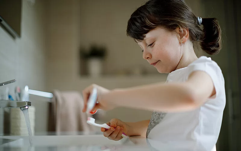 Ein etwa 7-jähriges Mädchen steht an einem Waschbecken, bei dem der Wasserhahn aufgedreht ist. Sie ist im Begriff, Zahnpasta aus einer Tube auf ihre Zahnbürste zu drücken.