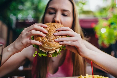 Eine junge blonde Frau hält einen fetten Burger in den Händen und beißt hinein.