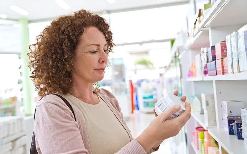 Eine Frau mittleren Alters mit roten Locken steht in einer Drogerie und liest die Liste der Inhaltstoffe auf einer Cremedose.