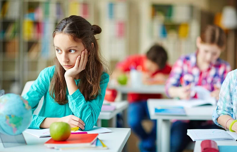 Eine circa 8-jährige Schülerin sitzt an ihrem Platz in der Klasse und schaut verträumt zur Seite, während alle anderen konzentriert arbeiten.