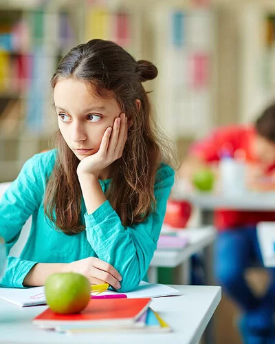 Eine circa 8-jährige Schülerin sitzt an ihrem Platz in der Klasse und schaut verträumt zur Seite, während alle anderen konzentriert arbeiten.