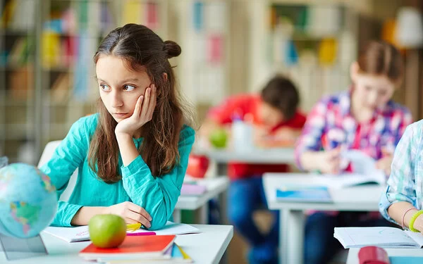 Eine circa 8-jährige Schülerin sitzt an ihrem Platz in der Klasse und schaut verträumt zur Seite, während alle anderen konzentriert arbeiten.