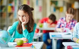 Eine circa 8-jährige Schülerin sitzt an ihrem Platz in der Klasse und schaut verträumt zur Seite, während alle anderen konzentriert arbeiten.