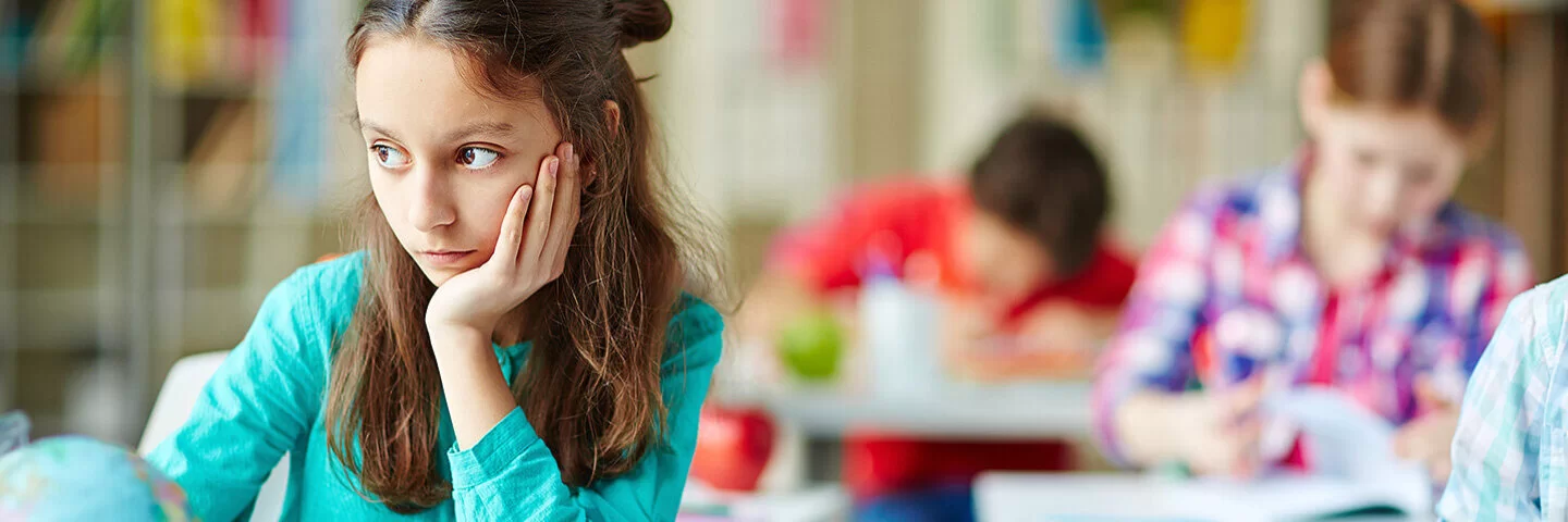 Eine circa 8-jährige Schülerin sitzt an ihrem Platz in der Klasse und schaut verträumt zur Seite, während alle anderen konzentriert arbeiten.