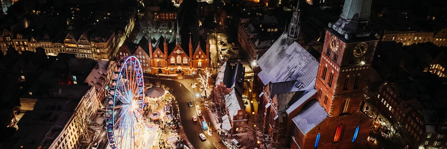 Nachtansicht aus der Vogelperspektive auf den belebten maritimen Weihnachtsmarkt auf dem Koberg in Lübeck. Ein großes, beleuchtetes Riesenrad dominiert die Szene, umgeben von Marktständen und festlicher Beleuchtung. Die umliegenden gotischen Gebäude, die St. Petri Kirche und das Heiligen-Geist-Hospital, sind festlich erleuchtet, was die weihnachtliche Stimmung der Szene unterstreicht. Die Dächer der Gebäude sind leicht mit Schnee bedeckt.