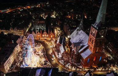 Nachtansicht aus der Vogelperspektive auf den belebten maritimen Weihnachtsmarkt auf dem Koberg in Lübeck. Ein großes, beleuchtetes Riesenrad dominiert die Szene, umgeben von Marktständen und festlicher Beleuchtung. Die umliegenden gotischen Gebäude, die St. Petri Kirche und das Heiligen-Geist-Hospital, sind festlich erleuchtet, was die weihnachtliche Stimmung der Szene unterstreicht. Die Dächer der Gebäude sind leicht mit Schnee bedeckt.