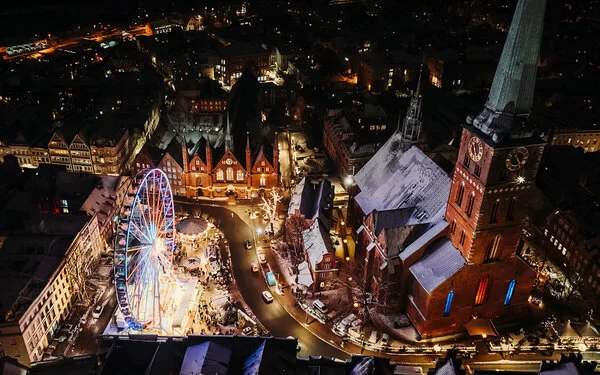 Nachtansicht aus der Vogelperspektive auf den belebten maritimen Weihnachtsmarkt auf dem Koberg in Lübeck. Ein großes, beleuchtetes Riesenrad dominiert die Szene, umgeben von Marktständen und festlicher Beleuchtung. Die umliegenden gotischen Gebäude, die St. Petri Kirche und das Heiligen-Geist-Hospital, sind festlich erleuchtet, was die weihnachtliche Stimmung der Szene unterstreicht. Die Dächer der Gebäude sind leicht mit Schnee bedeckt.