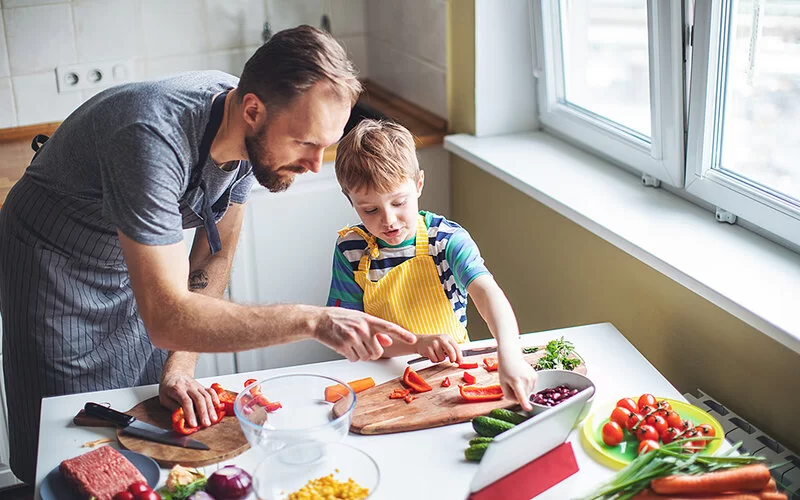 Ein Vater mit seinem ungefähr fünfjährigen Sohn in der Küche. Beide tragen Kochschürzen und schneiden buntes Gemüse.