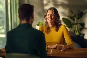 Ein nicht erkennbarer Mann mit kurzen dunklen Haaren und dunklem Hemd sitzt an einem Holztisch einer Frau mit gewelltem, dunkelblonden Haar in einem gelben Pullover gegenüber. Auf dem Tisch stehen zwei Gläser mit Wasser, im Hintergrund sind Grünpflanzen zu sehen.