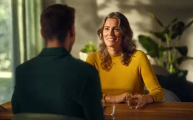 Ein nicht erkennbarer Mann mit kurzen dunklen Haaren und dunklem Hemd sitzt an einem Holztisch einer Frau mit gewelltem, dunkelblonden Haar in einem gelben Pullover gegenüber. Auf dem Tisch stehen zwei Gläser mit Wasser, im Hintergrund sind Grünpflanzen zu sehen.