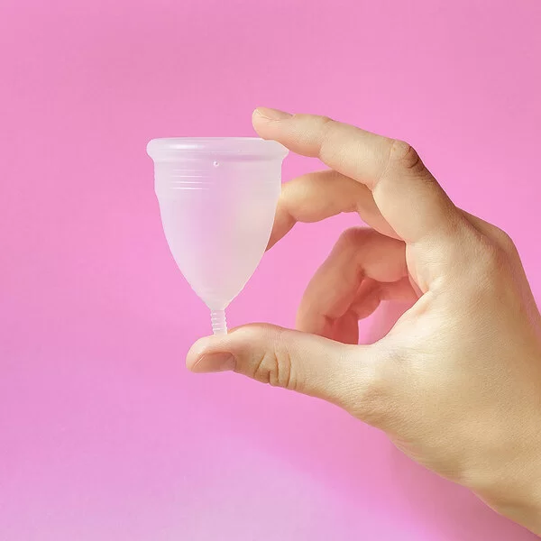 Eine weibliche Hand hält eine Menstruationstasse vor rosa Hintergrund. 