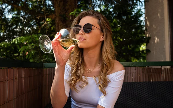 Eine junge Frau mit Sonnenbrille sitzt auf einem Balkon und trinkt Weißwein aus einem Weinglas.