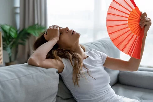 Eine Frau leidet unter Kreislaufproblemen durch Hitze und sitzt erschöpft auf einem Sofa, während sie sich Luft mit einem Fächer zu wedelt.