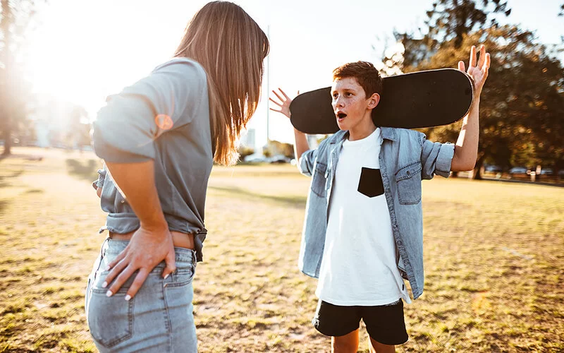 Eine Mutter, die man von hinten sieht, schimpft in einem Park mit ihrem Sohn, der vor ihr steht und sich sein Skateboard hinter den Kopf hält.