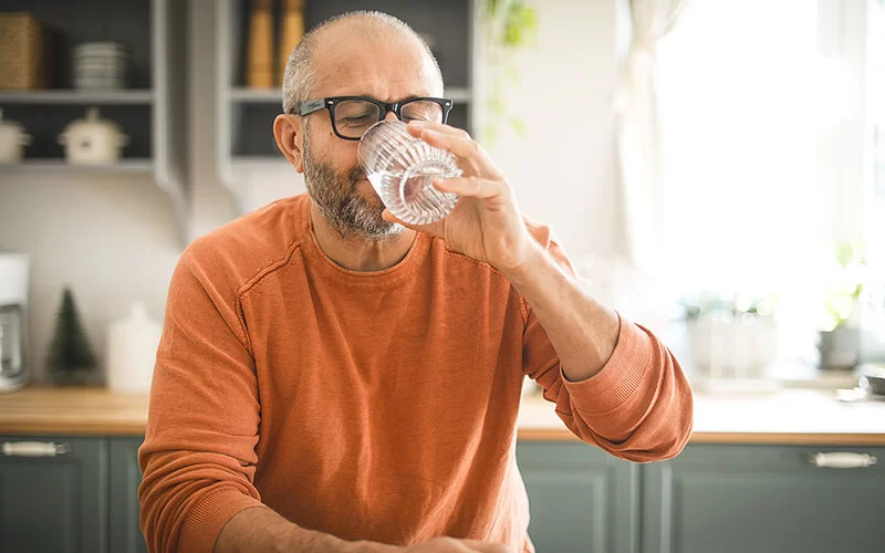 Ein älterer Mann mit Vollbart und dunkler Brille sitzt an einem Küchentisch und trinkt genussvoll ein Glas Wasser.