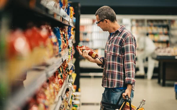 Ein Mann steht mit einem Einkaufskorb in einem Supermarkt vor einem Regal mit Knabberartikeln. Er hält eine Tüte Chips in der Hand und liest die Informationen auf der Verpackung.