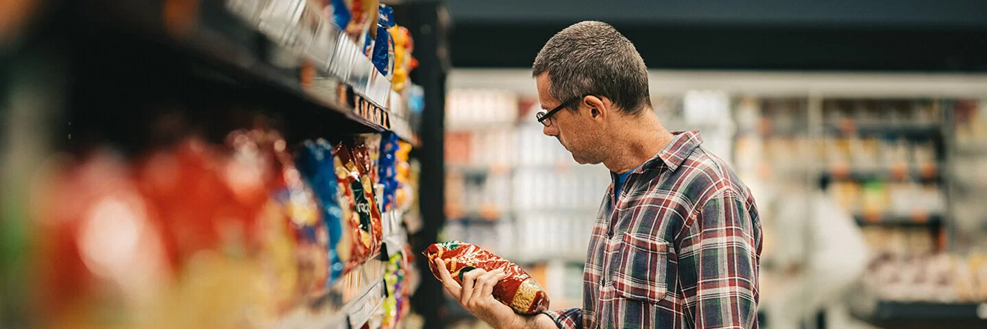 Ein Mann steht mit einem Einkaufskorb in einem Supermarkt vor einem Regal mit Knabberartikeln. Er hält eine Tüte Chips in der Hand und liest die Informationen auf der Verpackung.