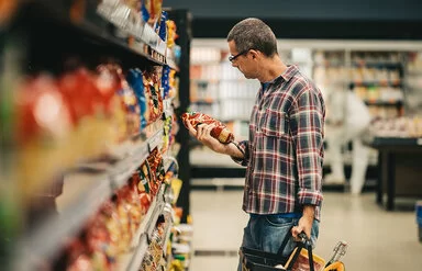 Ein Mann steht mit einem Einkaufskorb in einem Supermarkt vor einem Regal mit Knabberartikeln. Er hält eine Tüte Chips in der Hand und liest die Informationen auf der Verpackung.