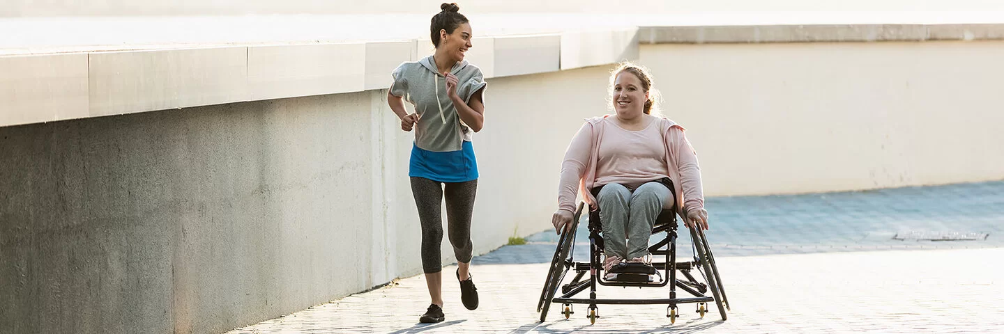 Zwei lachende junge Frauen an einer Uferpromenade. Eine der beiden sitzt im Rollstuhl, die andere joggt neben ihr her.