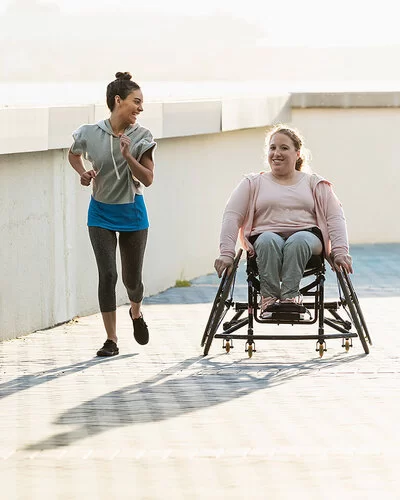 Zwei lachende junge Frauen an einer Uferpromenade. Eine der beiden sitzt im Rollstuhl, die andere joggt neben ihr her.