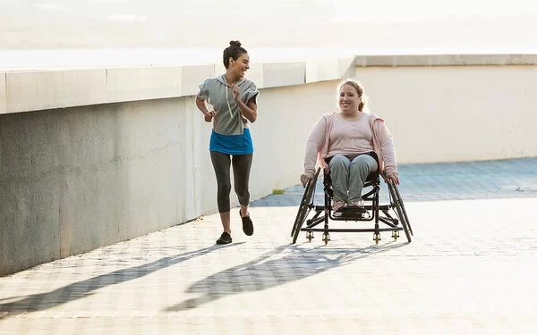 Zwei lachende junge Frauen an einer Uferpromenade. Eine der beiden sitzt im Rollstuhl, die andere joggt neben ihr her.