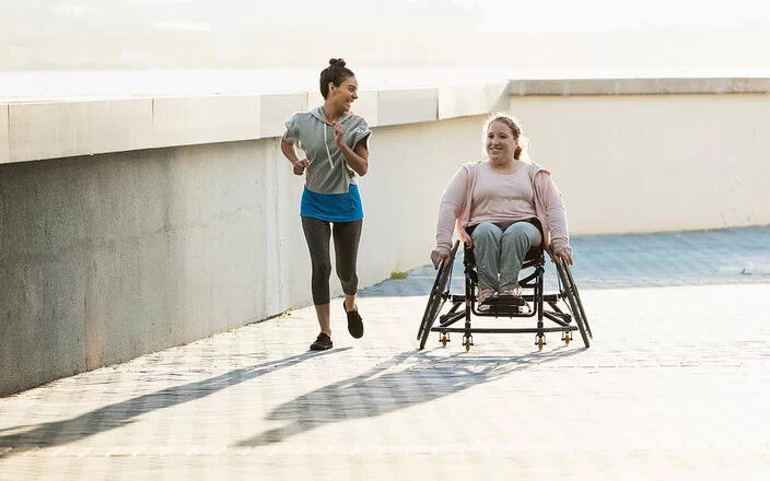 Zwei lachende junge Frauen an einer Uferpromenade. Eine der beiden sitzt im Rollstuhl, die andere joggt neben ihr her.