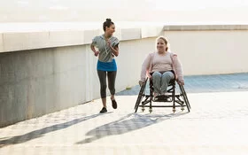 Zwei lachende junge Frauen an einer Uferpromenade. Eine der beiden sitzt im Rollstuhl, die andere joggt neben ihr her.