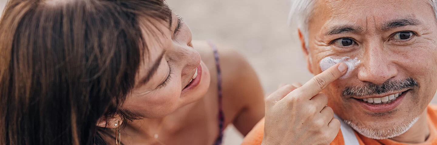Eine Frau cremt einem Mann das Gesicht mit Sonnencreme ein.