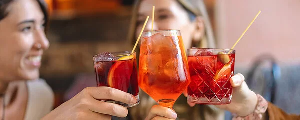 Zwei Frauen stoßen lächelnd mit alkoholischen Getränken ein. Eine der beiden hält einen orangefarbenen Aperol Spritz in der Hand, die andere einen roten Longdrink. Rechts ist ein weiteres Glas mit rotem Inhalt zu sehen, das ebenfalls zum Anstoßen erhoben ist.