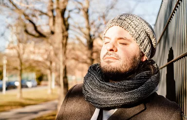 Ein Mann in herbstlicher Kleidung lehnt sich an einen Zaun, die Sonne scheint ihm ins Gesicht.