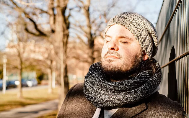 Ein Mann in herbstlicher Kleidung lehnt sich an einen Zaun, die Sonne scheint ihm ins Gesicht.