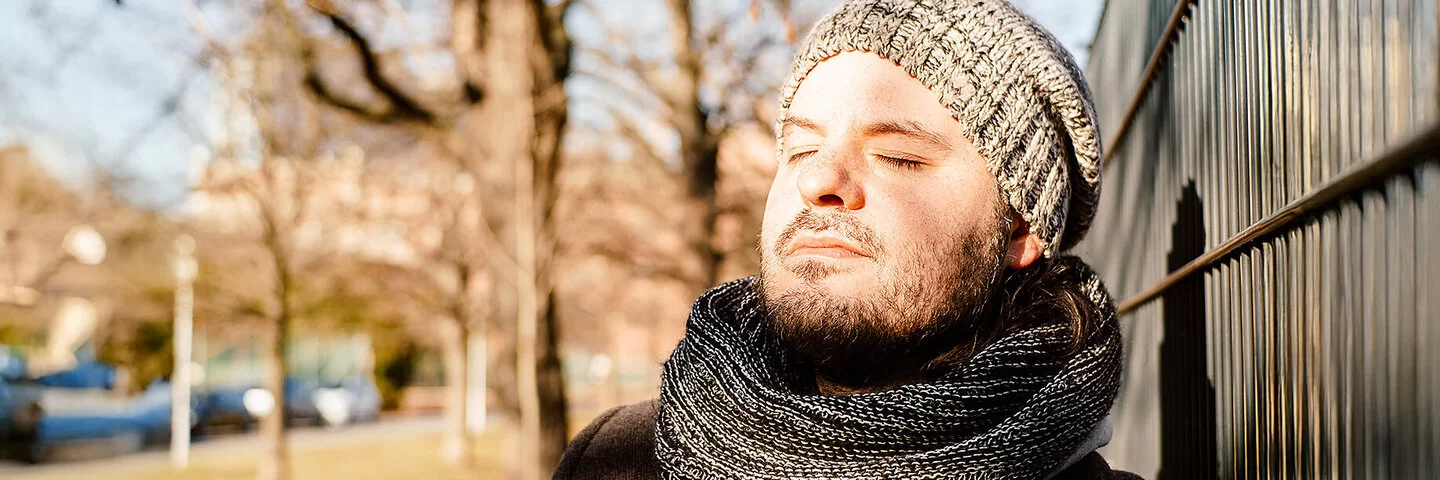 Ein Mann in herbstlicher Kleidung lehnt sich an einen Zaun, die Sonne scheint ihm ins Gesicht.