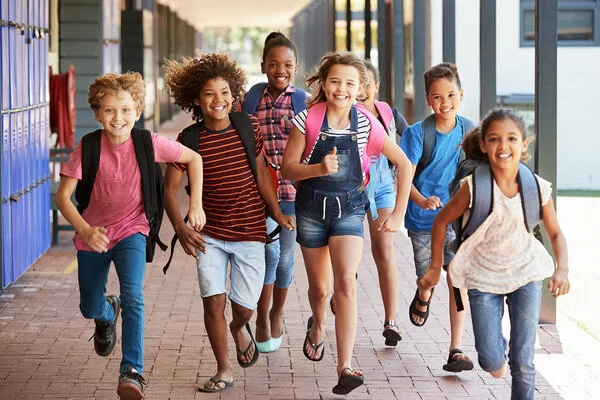 Sieben junge Schüler, vier Mädchen, drei Jungs- mit unterschiedlichen Hautfarben, bunt gekleidet, rennen strahlend mit ihren Schulrucksäcken durch eine Schule.