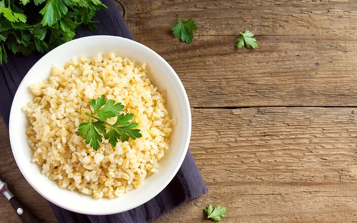 Eine weiße Schale ist gefüllt mit Bulgur aus Weizenkörnern. Dekoriert ist er mit frischer Petersilie. Neben der Schale liegen noch einzelne Blätter und Reste der Petersilie. Die Schale steht auf einem lilafarbenen Tischset, das auf einer dunklen, rustikalen Holzplatte liegt.